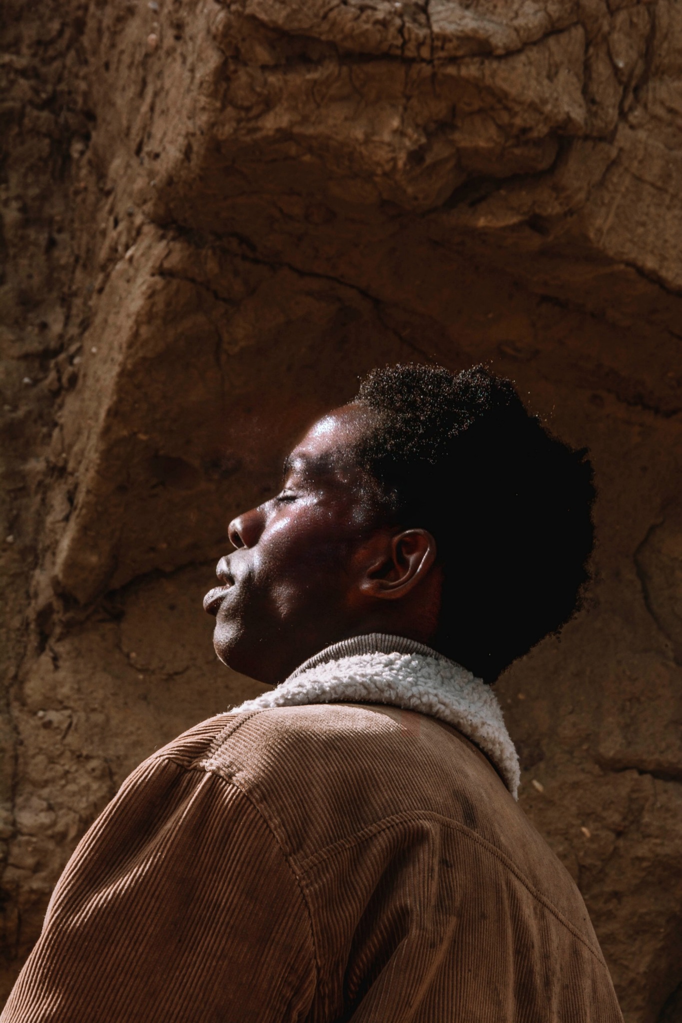 Portret z profilu, mężczyzna z ciemną karnacją, w brązowej kurtce sztruksowej z jasnym kołnierzem z baranka, na tle skalnej ściany oświetlony ciepłym światłem.