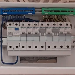 Wnętrze skrzynki elektrycznej z zainstalowanymi bezpiecznikami, widoczne niebieskie i zielone listwy zaciskowe oraz okablowanie.