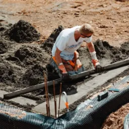 Pracownik w okularach i rękawicach wyrównuje betonową wylewkę fundamentu za pomocą długiej poziomicy, widoczne zbrojenie i folia kubełkowa.