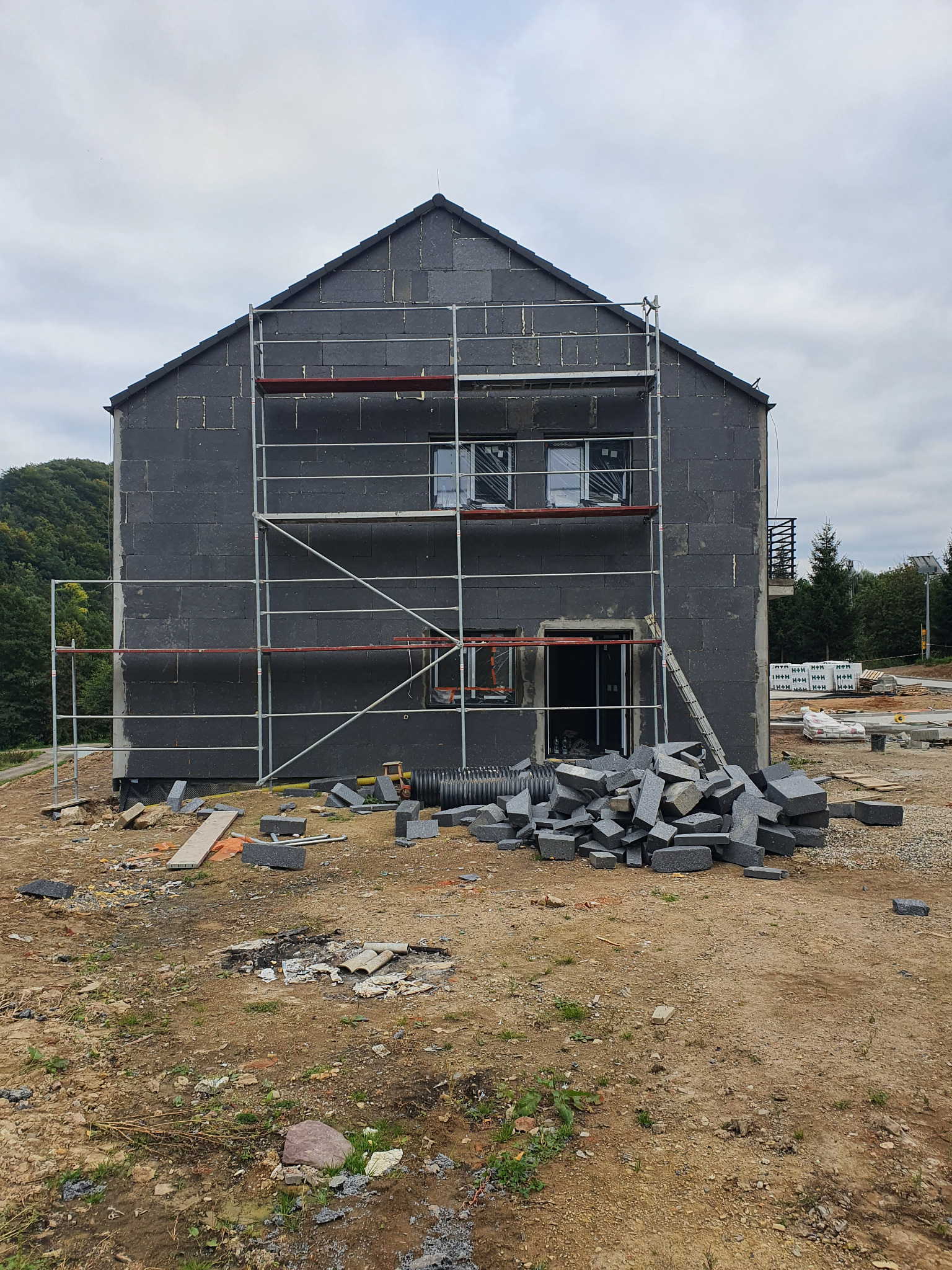 Surowy stan domu jednorodzinnego z szarą elewacją i rusztowaniami, przygotowany do dalszych prac wykończeniowych, z widocznym stosem bloczków.