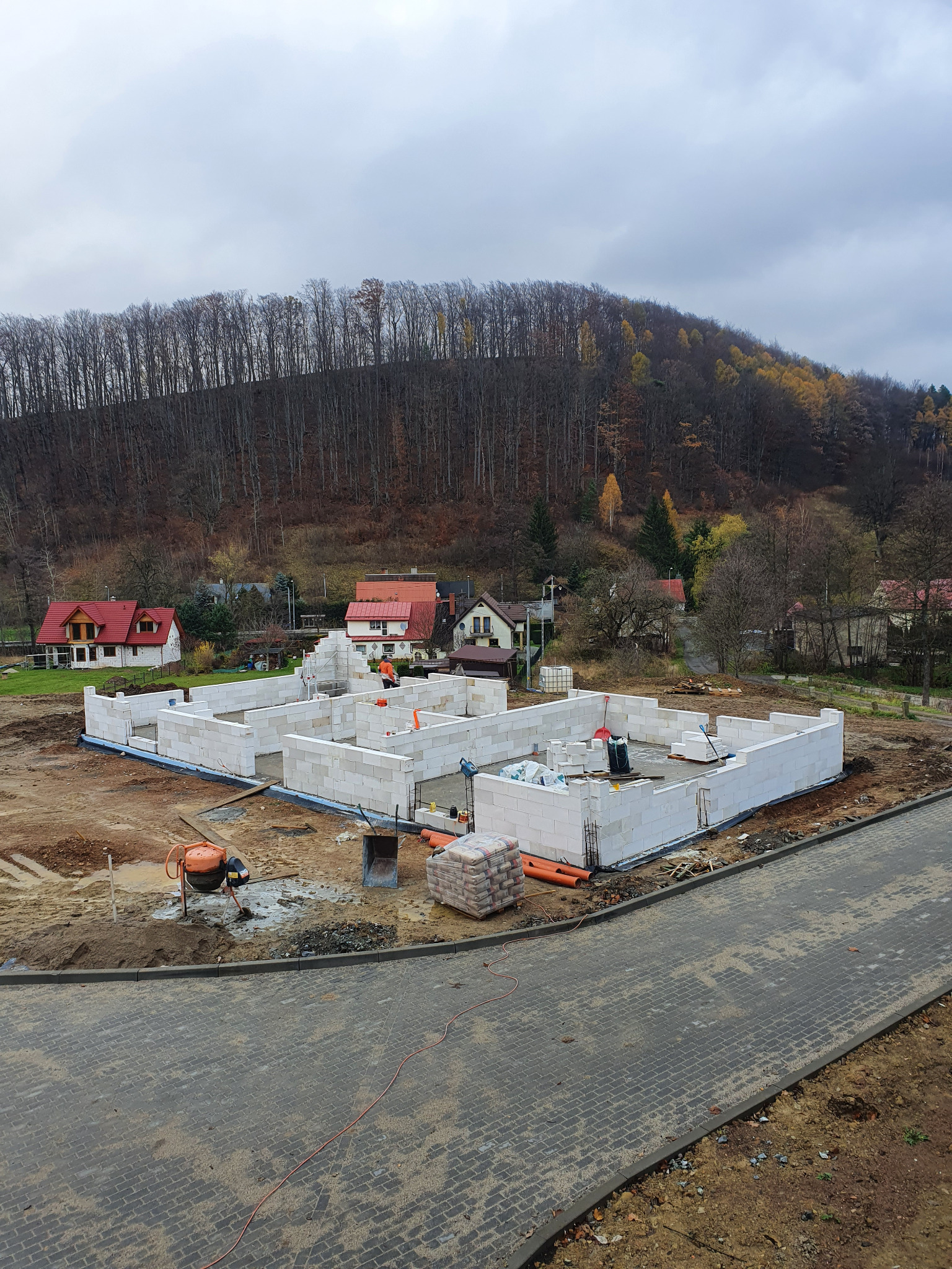 Budowa domu z białych bloczków: widok z góry na fundamenty i ściany parteru na tle jesiennego krajobrazu z widocznymi domami w oddali.