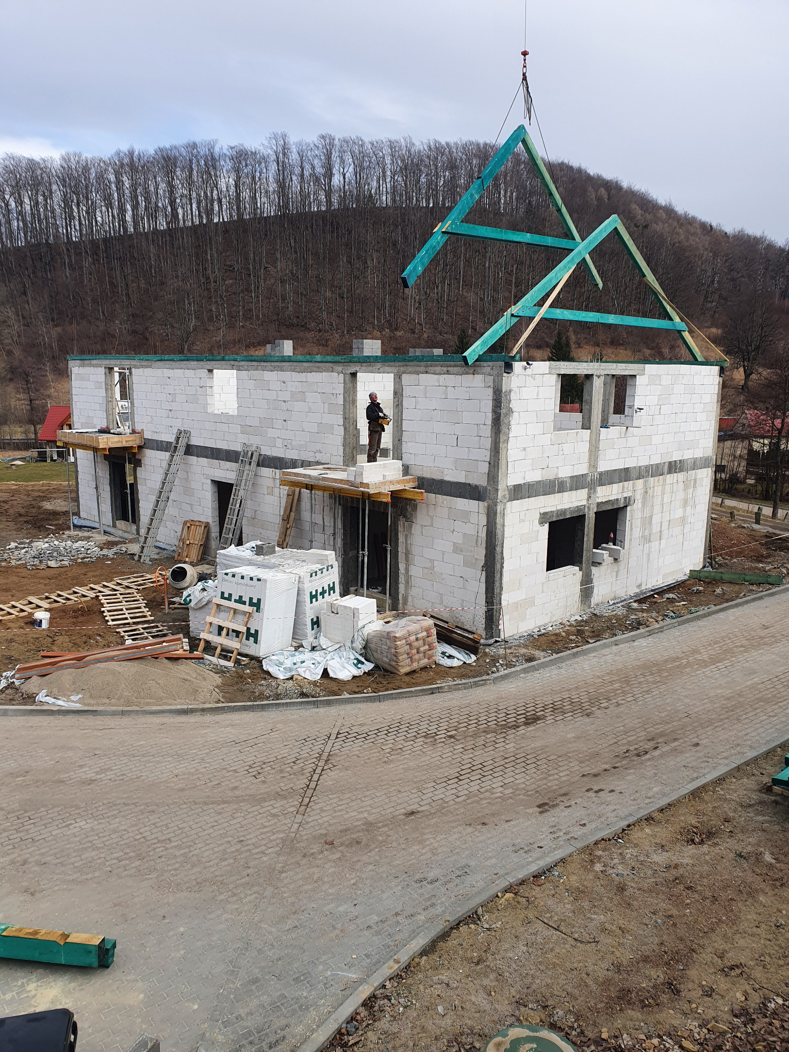 Wznoszenie szczytu dachu z belek pomalowanych na zielono na dwukondygnacyjnym domu z białych bloczków, widok na wzgórze z drzewami.