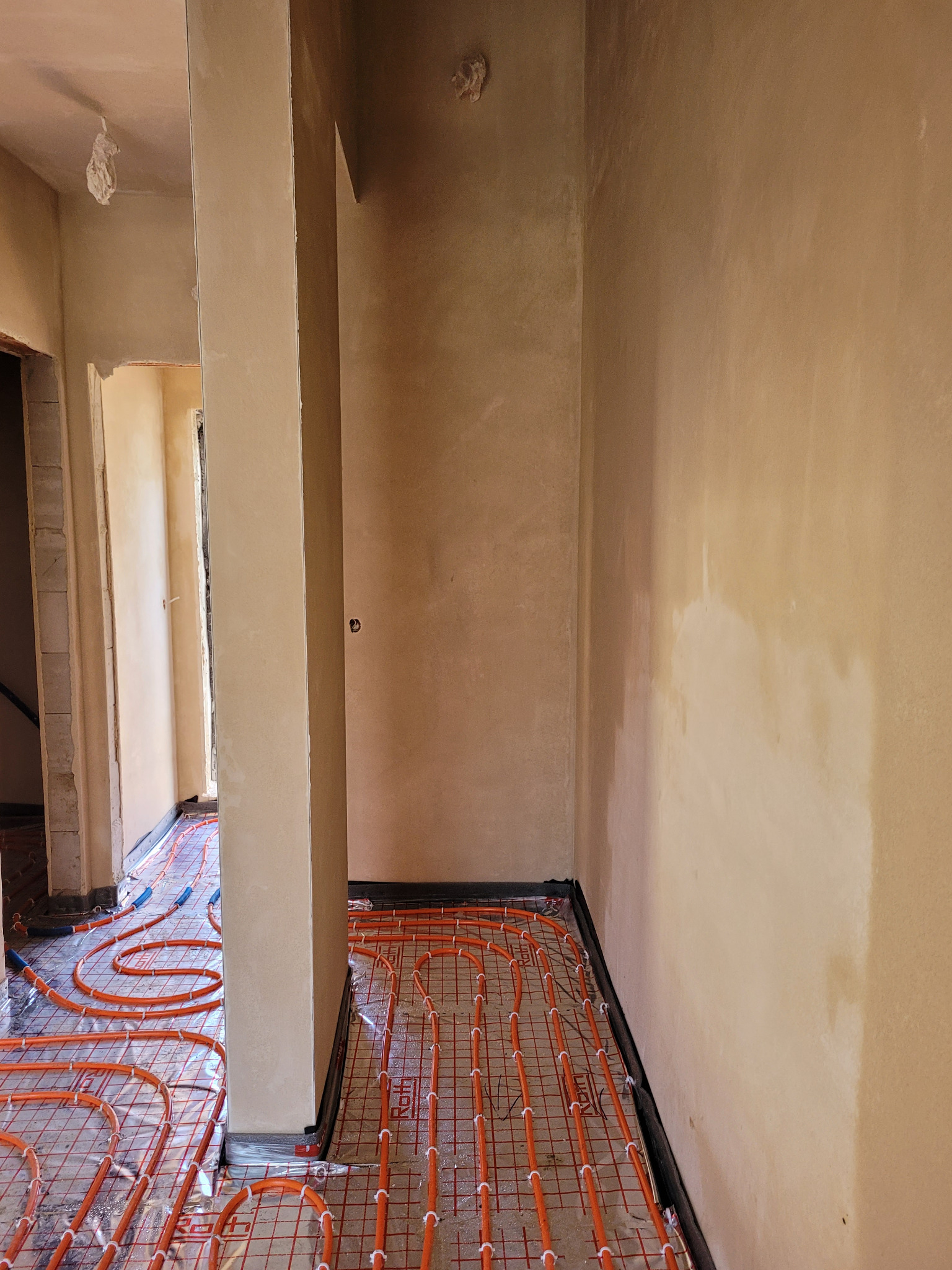Wnętrze w trakcie prac wykończeniowych: ściany otynkowane na beżowo, na podłodze rozłożona instalacja ogrzewania podłogowego z pomarańczowych rurek ułożonych w regularną siatkę, zabezpieczona folią.