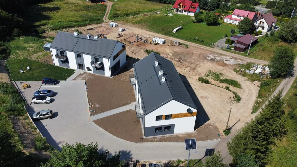 Dwa nowoczesne budynki wielorodzinne z szarymi dachami i białymi elewacjami, widok z lotu ptaka, w tle plac budowy z fundamentami i materiałami budowlanymi.