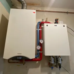 Biały, wiszący piec gazowy Termet z wyświetlaczem wskazującym 50 stopni Celsjusza, połączony rurami w kolorze czerwonym i niebieskim, z manometrami, zamontowany na surowej ścianie w trakcie...