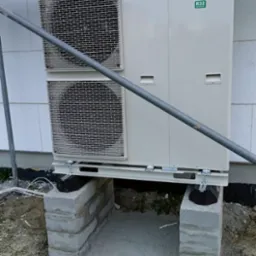 Zewnętrzna jednostka pompy ciepła Panasonic R32, zamontowana na betonowych blokach fundamentowych z wylewką, widoczna na tle ściany budynku w trakcie budowy.