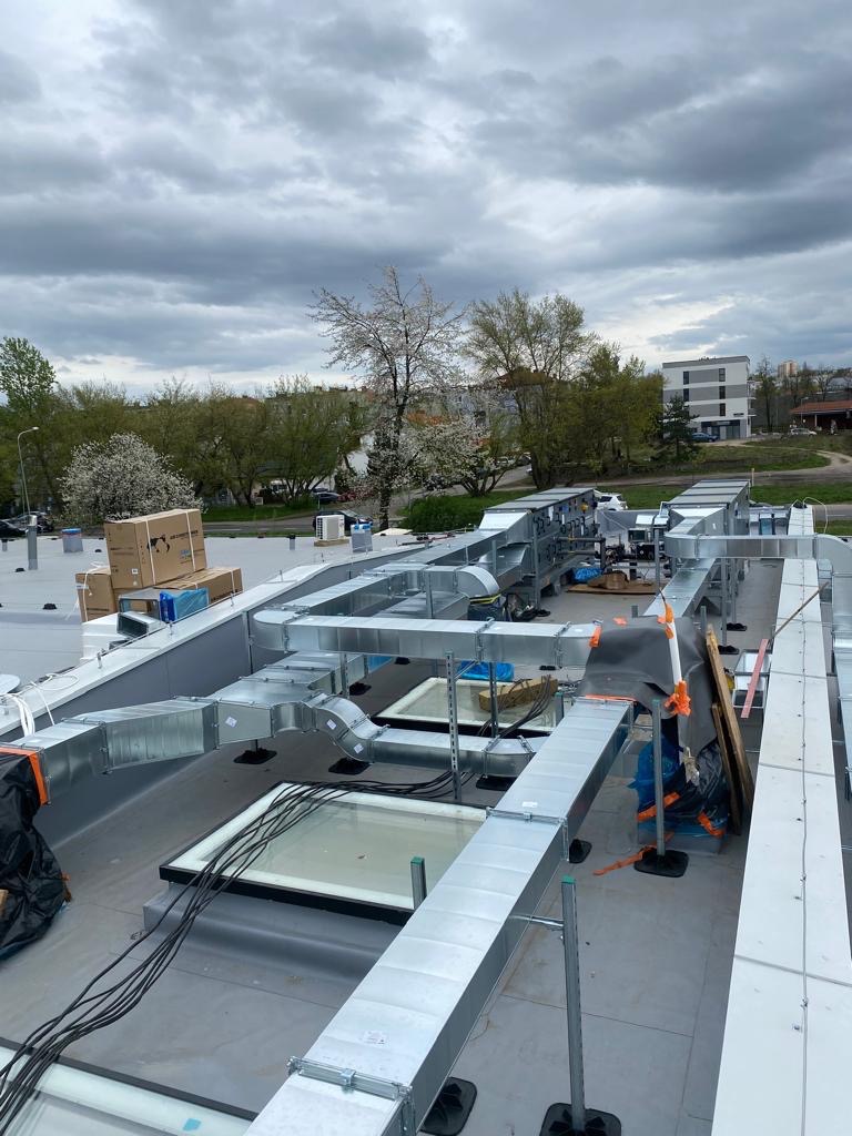 Widok z góry na montaż systemu kanałów wentylacyjnych na dachu budynku, z widocznymi elementami mocującymi, skrzyniami i przewodami, na tle pochmurnego nieba i zieleni miejskiej.