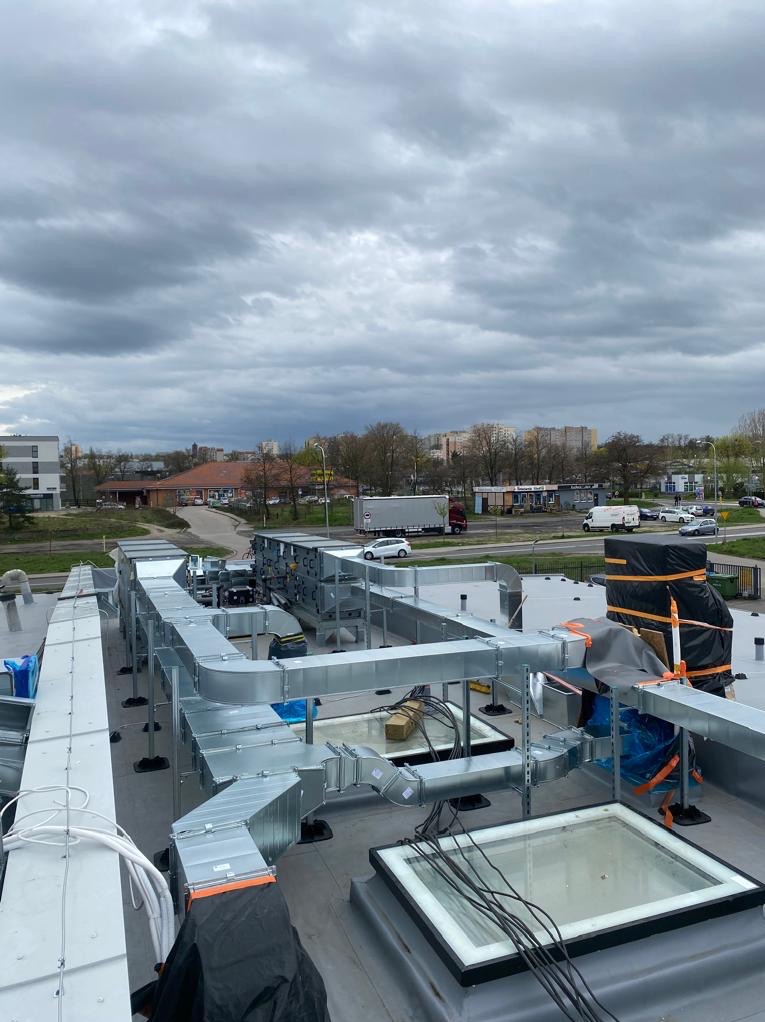 Kompleksowy system kanałów wentylacyjnych z blachy ocynkowanej na dachu budynku, widok z góry w pochmurny dzień, w tle zabudowa miejska i ruch uliczny.