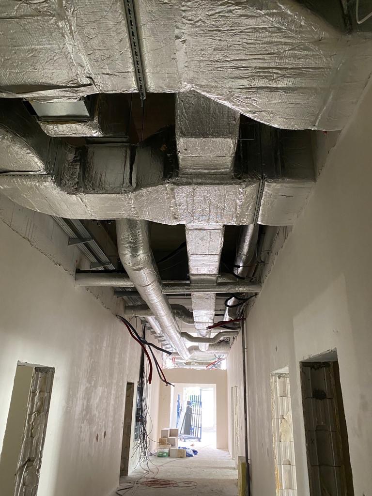 Widok z dołu na sufit z zamontowanymi, zaizolowanymi przewodami wentylacyjnymi w surowym wnętrzu budynku w trakcie wykańczania, z widocznymi kablami i otworami drzwiowymi.