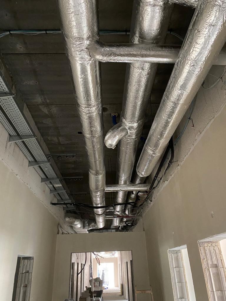 Srebrne, zaizolowane rury wentylacyjne pod sufitem w surowym stanie budynku, widoczne koryta kablowe i instalacje elektryczne.