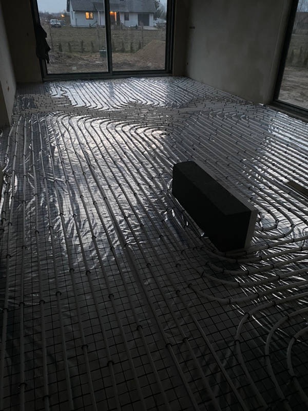 Rozłożona instalacja ogrzewania podłogowego na folii aluminiowej w surowym wnętrzu, widoczne rury grzewcze i siatka montażowa, w tle okno z widokiem na dom w budowie.