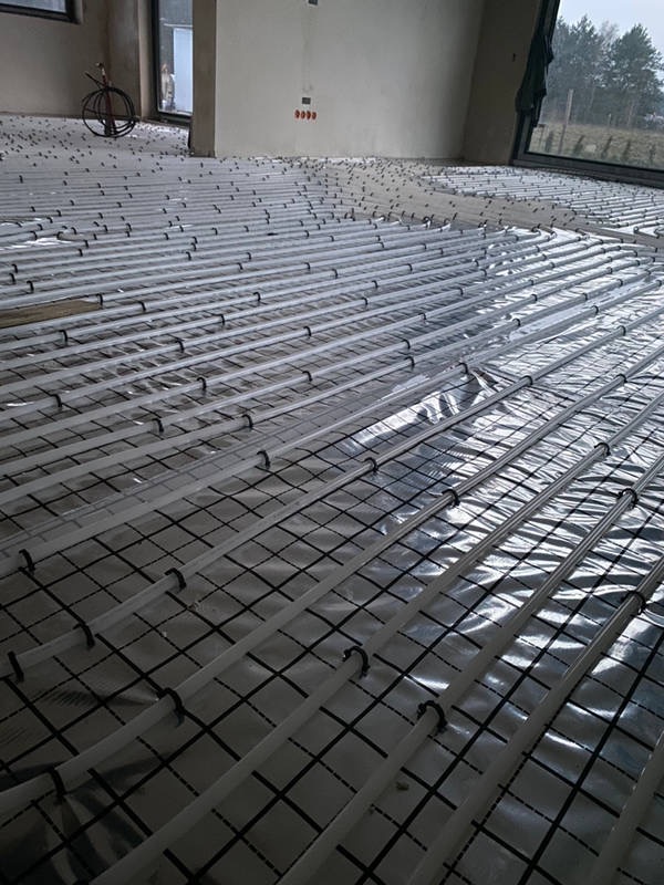 Białe rury ogrzewania podłogowego zamocowane na folii z siatką, widok z góry w surowym wnętrzu z dużym oknem.