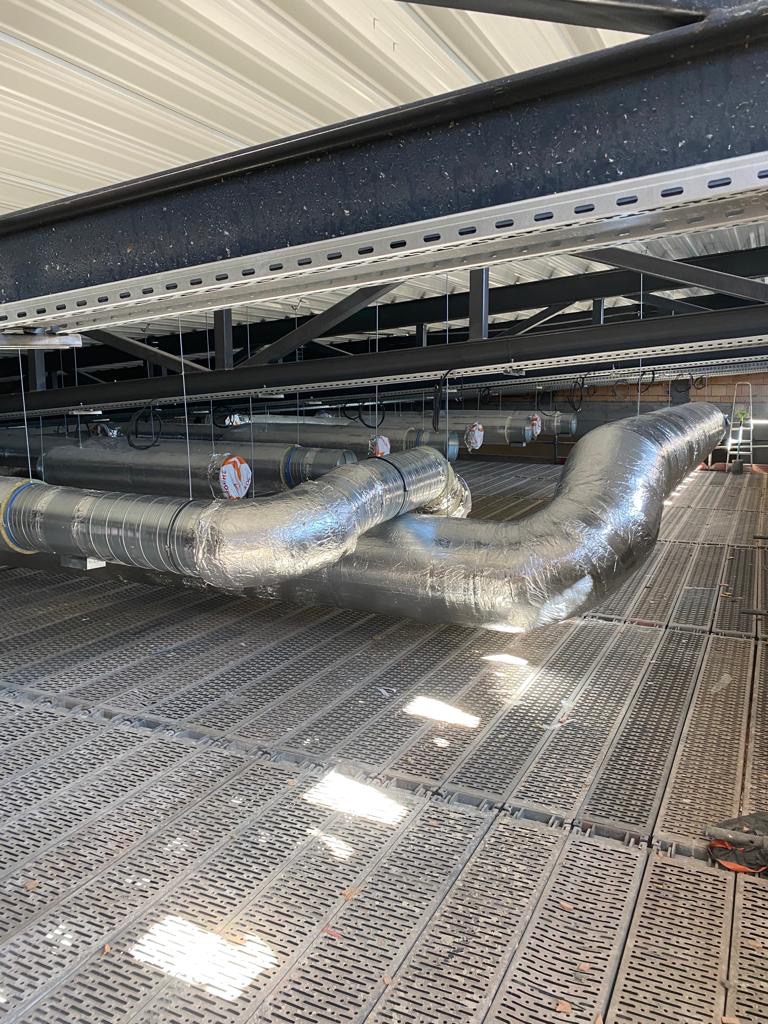 Srebrne, izolowane przewody wentylacyjne zamontowane pod sufitem w budynku przemysłowym, widoczne stalowe konstrukcje nośne i perforowana podłoga techniczna.