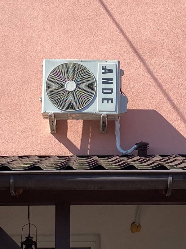 Biała jednostka zewnętrzna klimatyzacji marki ANDE zamontowana na różowej ścianie budynku, widoczna rura odprowadzająca skropliny nad zadaszeniem.