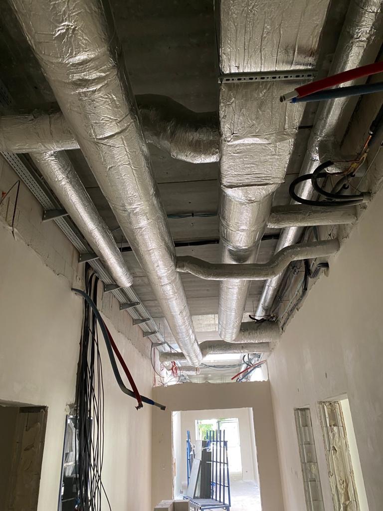 Widok sufitu w surowym stanie z licznymi, zaizolowanymi przewodami wentylacyjnymi i instalacyjnymi, metalowymi wspornikami i zwisającymi kablami.