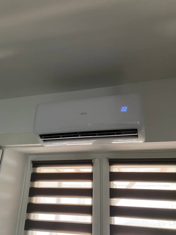 Biały klimatyzator ścienny z wyświetlaczem temperatury 22 stopnie Celsjusza, zamontowany nad oknem z roletami w odcieniach brązu.