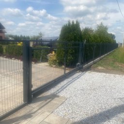 ArtSpawDesign MIODEK Marek Borowczak - Otwarta, przesuwna brama panelowa w kolorze grafitowym z automatyką, prowadząca na posesję z wysypanym jasnym żwirem, w tle dom jednorodzinny i rząd tuj.