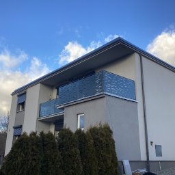 ArtSpawDesign MIODEK Marek Borowczak - Nowoczesny budynek mieszkalny z balkonami zabezpieczonymi ażurowymi, metalowymi balustradami w kolorze grafitowym, na tle błękitnego nieba z chmurami.