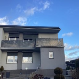 ArtSpawDesign MIODEK Marek Borowczak - Dwupiętrowy dom z szarą elewacją i balkonami z metalowymi balustradami o ażurowym wzorze, okna z roletami zewnętrznymi, widok na błękitne niebo z chmurami.