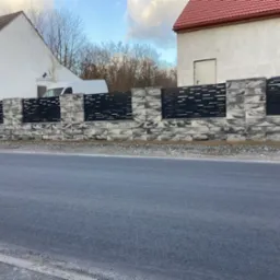 Nowoczesne ogrodzenie z prefabrykatów betonowych w odcieniach szarości i czarnych paneli, widok z poziomu ulicy, w tle budynki mieszkalne i zaparkowany biały bus.