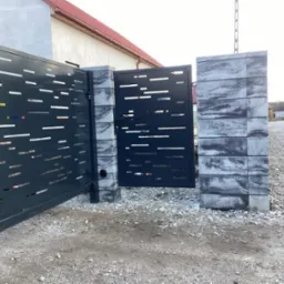 Nowoczesna brama wjazdowa przesuwna i furtka z metalu w kolorze antracytowym, z poziomymi ażurowymi wycięciami, osadzona na słupkach z betonowych bloczków imitujących kamień.