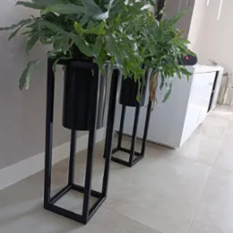 Dwa metalowe kwietniki w stylu loft z paprotkami na tle nowoczesnego salonu z białą komodą i szarą ścianą.