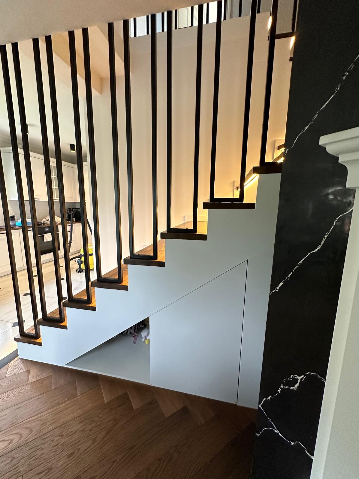 Nowoczesne schody wewnętrzne z drewnianymi stopniami i czarnymi, pionowymi balustradami, z ukrytą przestrzenią do przechowywania pod spodem i oświetleniem LED pod stopniami, na tle ściany z efektem...