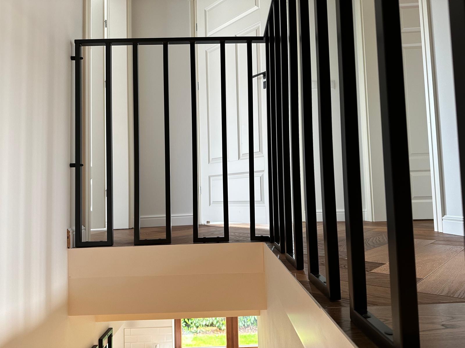 Nowoczesna, czarna, metalowa balustrada schodowa z pionowymi szczeblami w jasnym wnętrzu z drewnianą podłogą i białymi drzwiami.