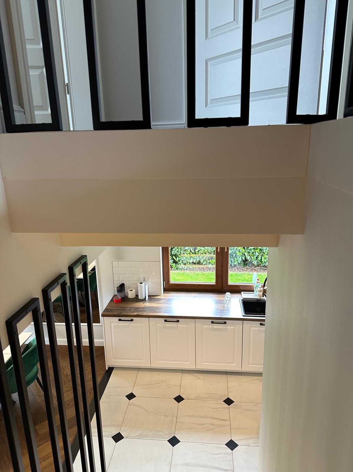 Widok z góry na białą kuchnię z drewnianym blatem i czarnymi akcentami, widzianą przez czarną metalową balustradę schodów.
