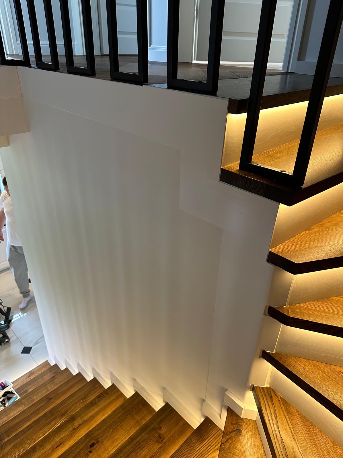 Nowoczesne schody drewniane z podświetleniem LED, czarne metalowe barierki, biała ściana, widok z góry.