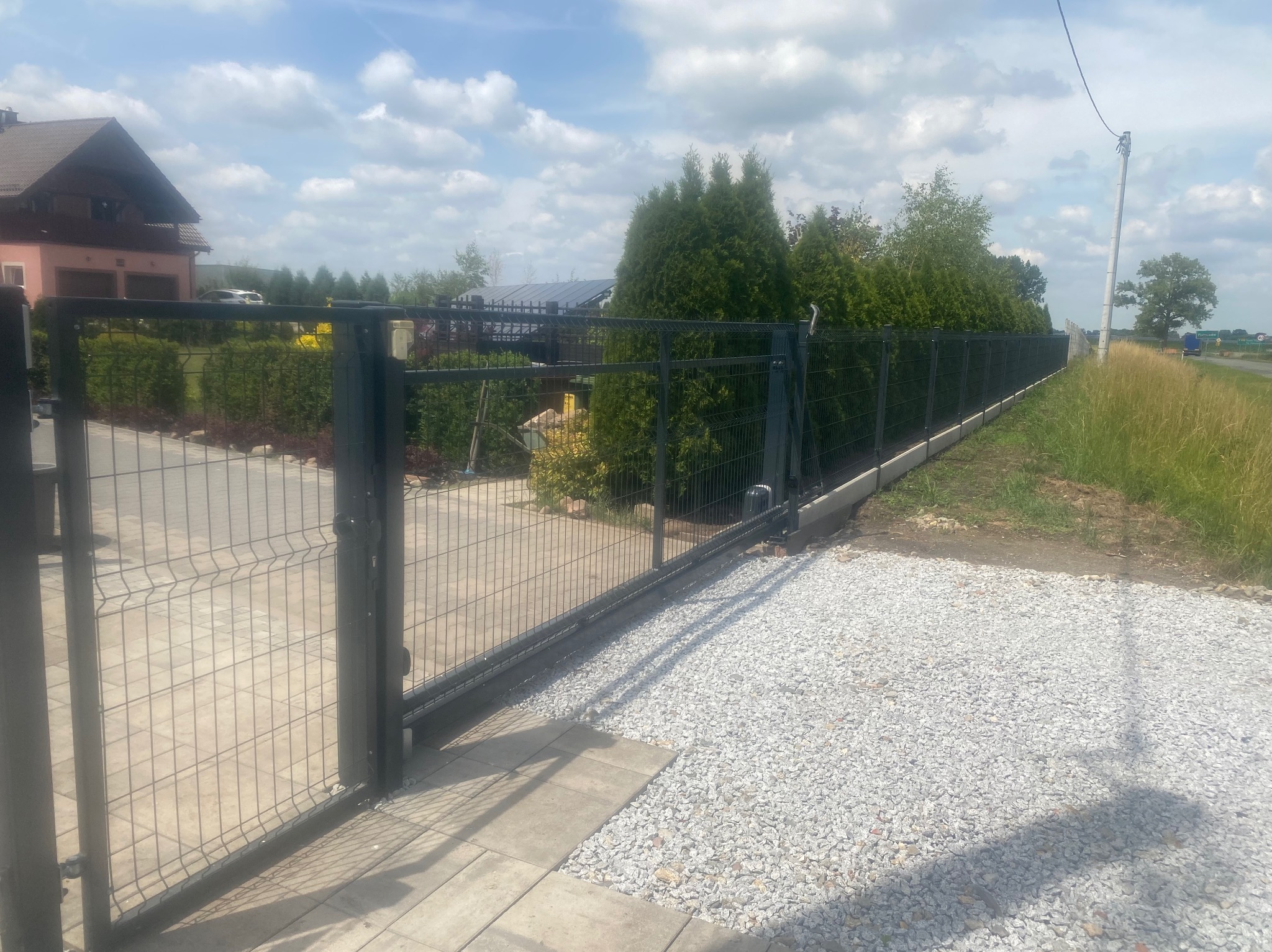 Otwarta, przesuwna brama panelowa w kolorze grafitowym z automatyką, prowadząca na posesję z wysypanym jasnym żwirem, w tle dom jednorodzinny i rząd tuj.
