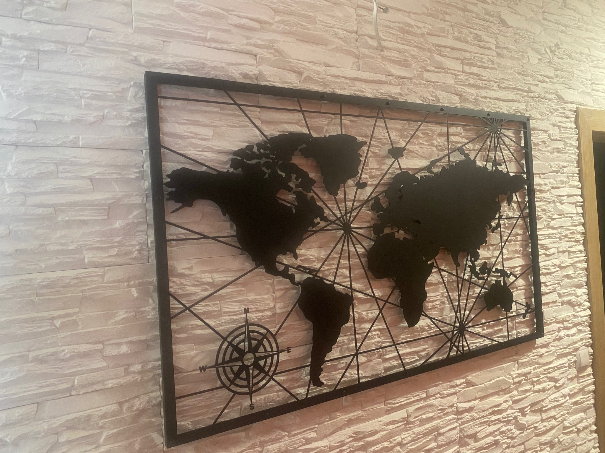 Metalowa mapa świata z kompasem, zawieszona na dekoracyjnej ścianie z białej cegły.