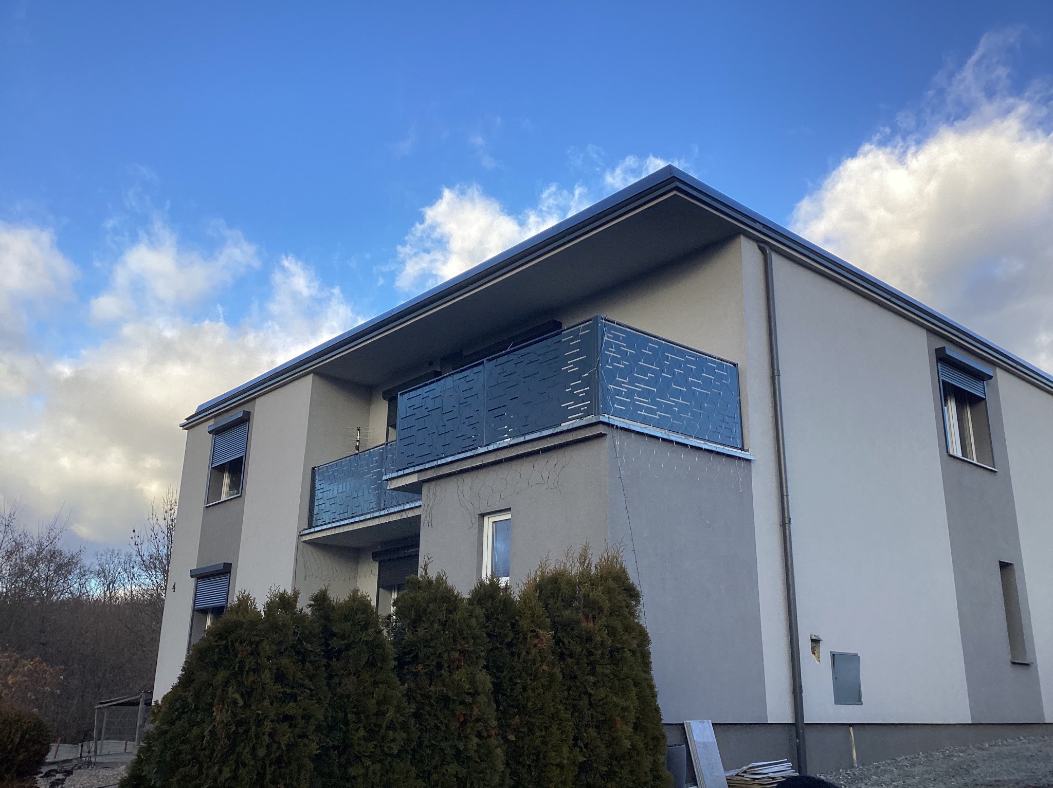 Nowoczesny budynek mieszkalny z balkonami zabezpieczonymi ażurowymi, metalowymi balustradami w kolorze grafitowym, na tle błękitnego nieba z chmurami.