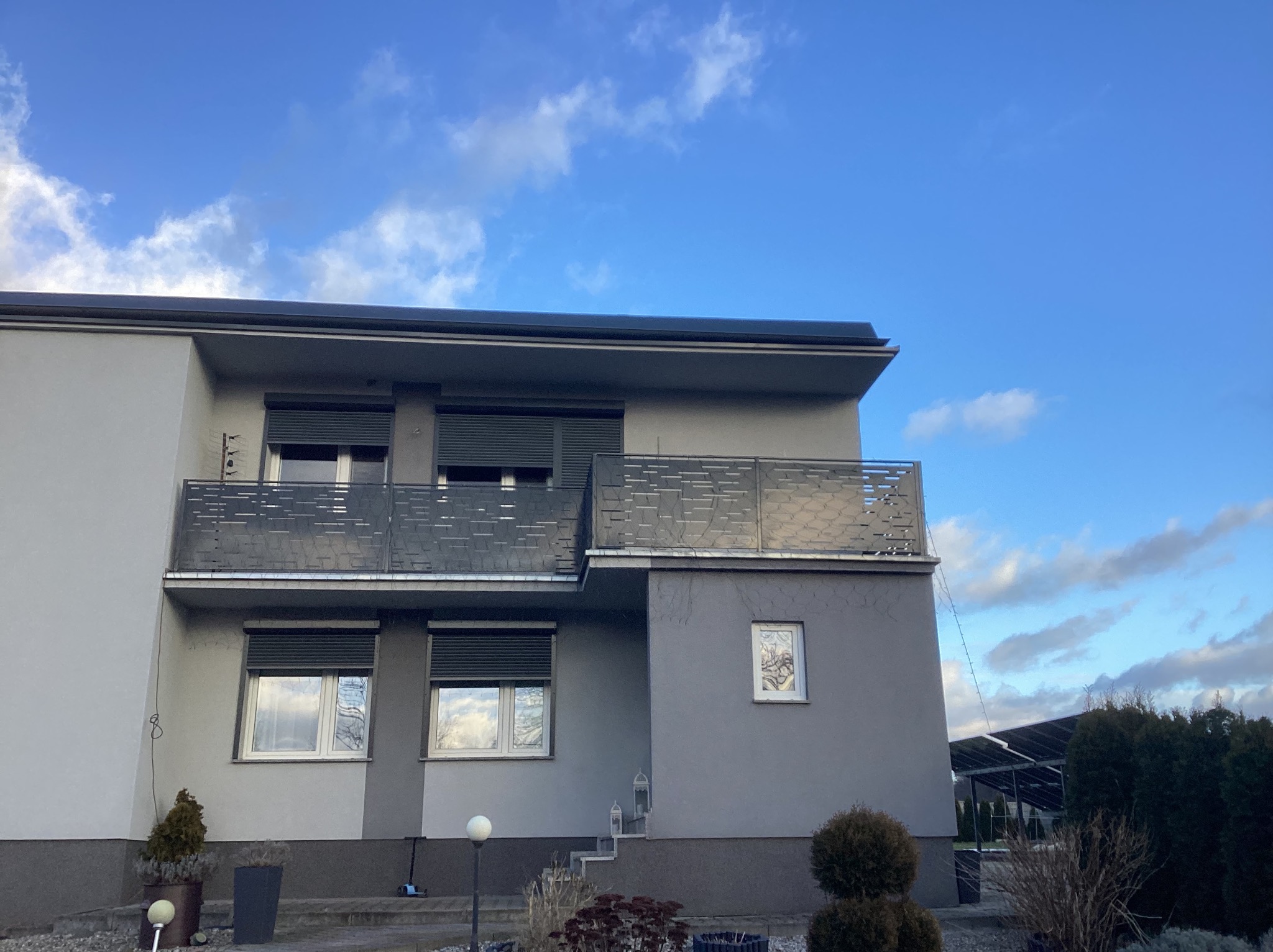 Dwupiętrowy dom z szarą elewacją i balkonami z metalowymi balustradami o ażurowym wzorze, okna z roletami zewnętrznymi, widok na błękitne niebo z chmurami.