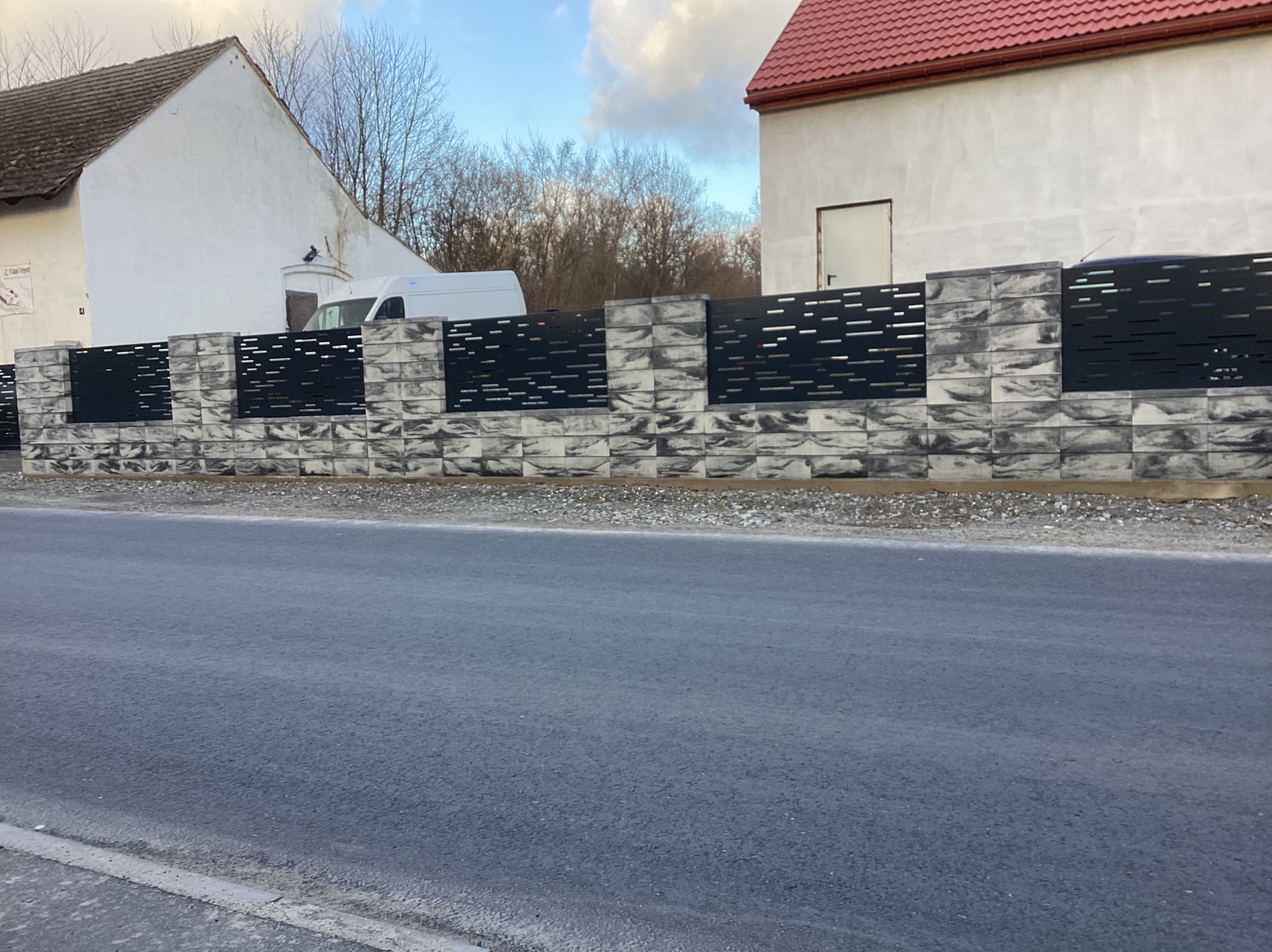 Nowoczesne ogrodzenie z prefabrykatów betonowych w odcieniach szarości i czarnych paneli, widok z poziomu ulicy, w tle budynki mieszkalne i zaparkowany biały bus.