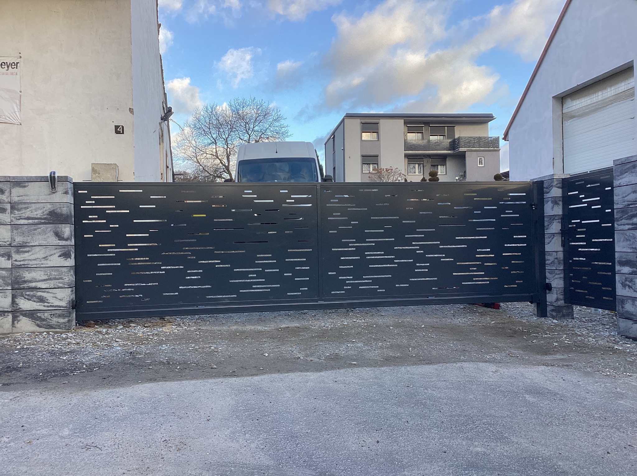 Nowoczesna, przesuwna brama wjazdowa z poziomymi, prostokątnymi wycięciami, zamontowana między dwoma budynkami, z widocznym białym vanem w tle.