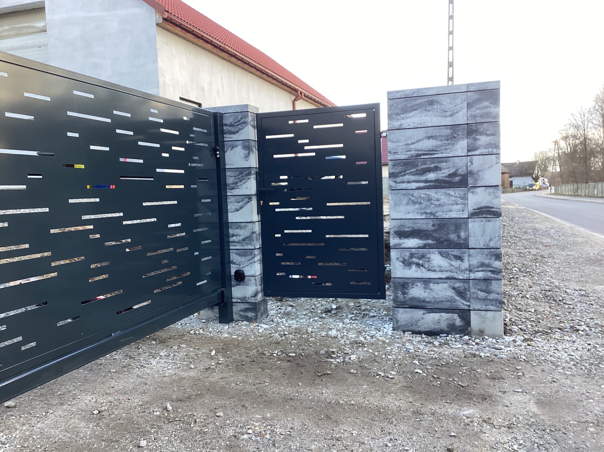 Nowoczesna brama wjazdowa przesuwna i furtka z metalu w kolorze antracytowym, z poziomymi ażurowymi wycięciami, osadzona na słupkach z betonowych bloczków imitujących kamień.