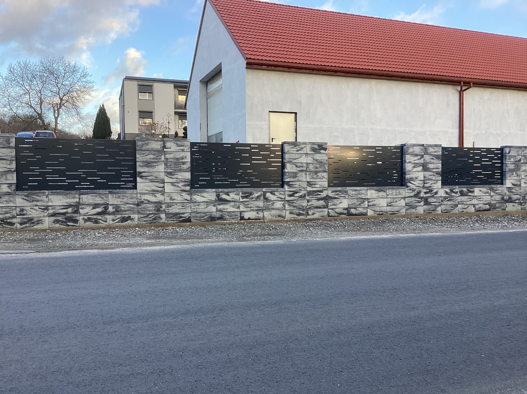 Nowoczesne ogrodzenie z szarego betonu architektonicznego i czarnych paneli z poziomymi wycięciami, widok z poziomu ulicy, w tle budynki i drzewa.