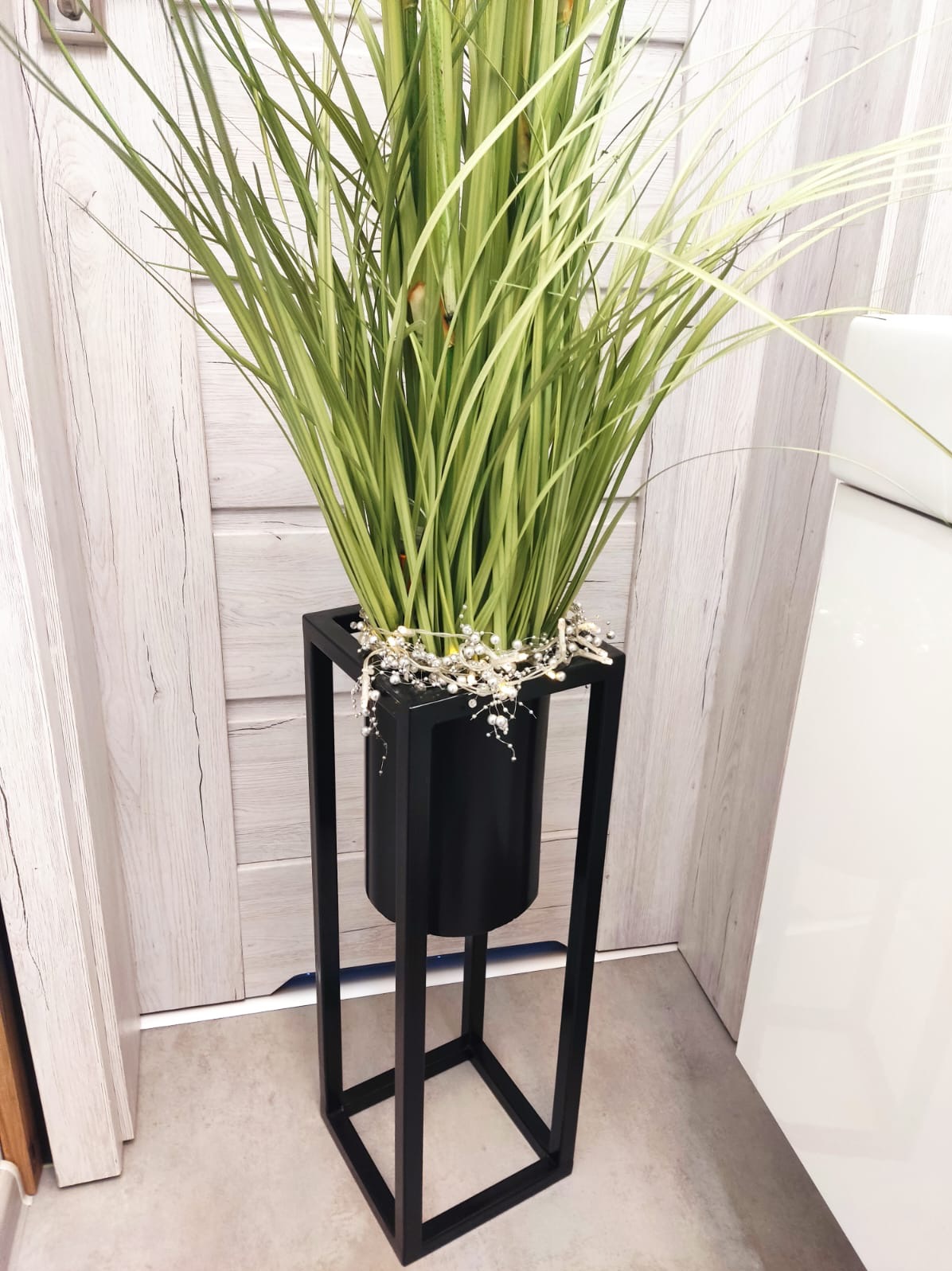Dekoracyjna, wysoka, czarna, metalowa podstawa na donicę z zielonymi trawami i drobnymi, białymi ozdobami, ustawiona w rogu pomieszczenia przy drewnianych drzwiach.