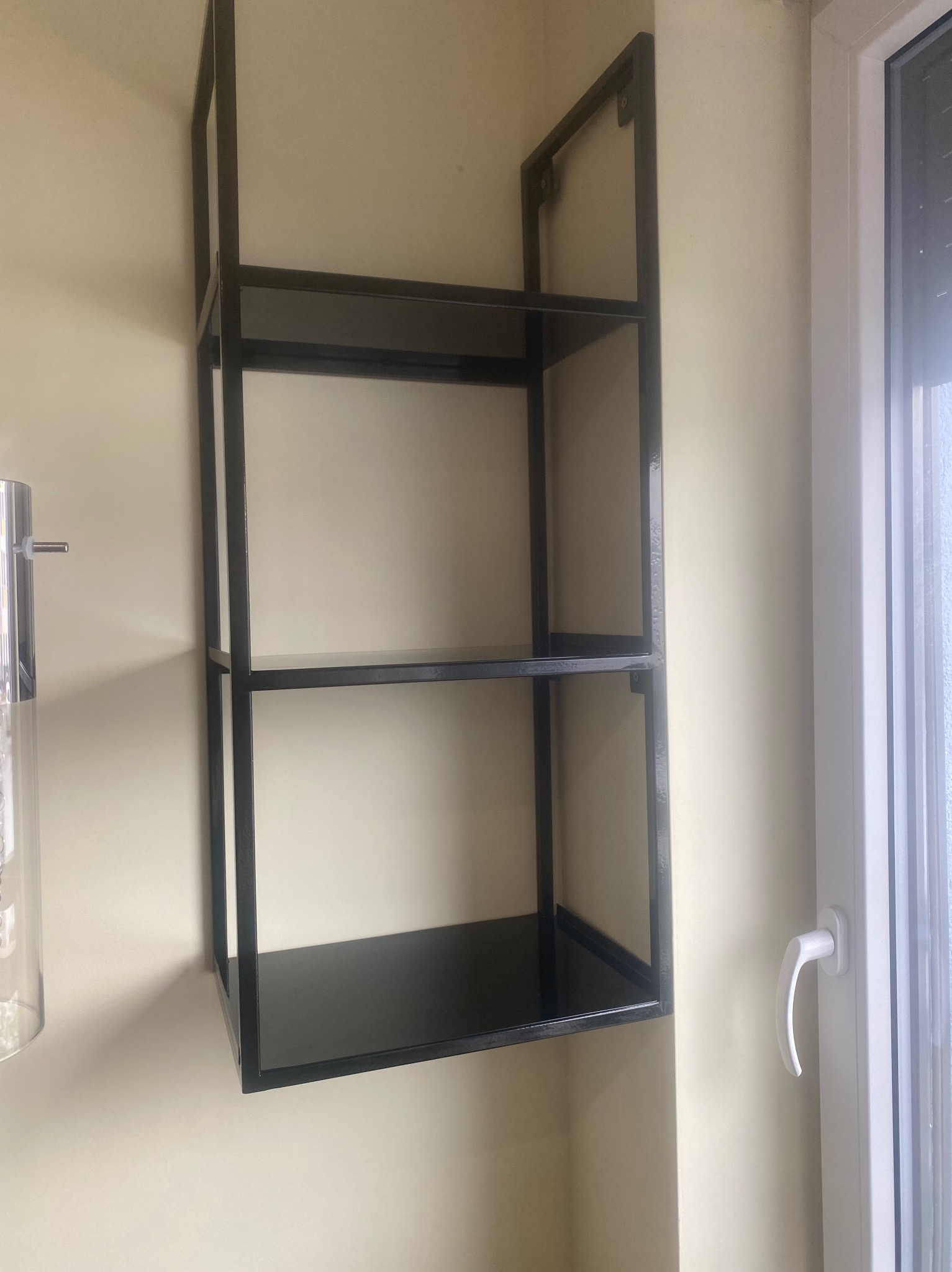 Metalowa, minimalistyczna półka ścienna z trzema poziomami, pomalowana na czarno, zamontowana na beżowej ścianie obok białego okna z roletą.