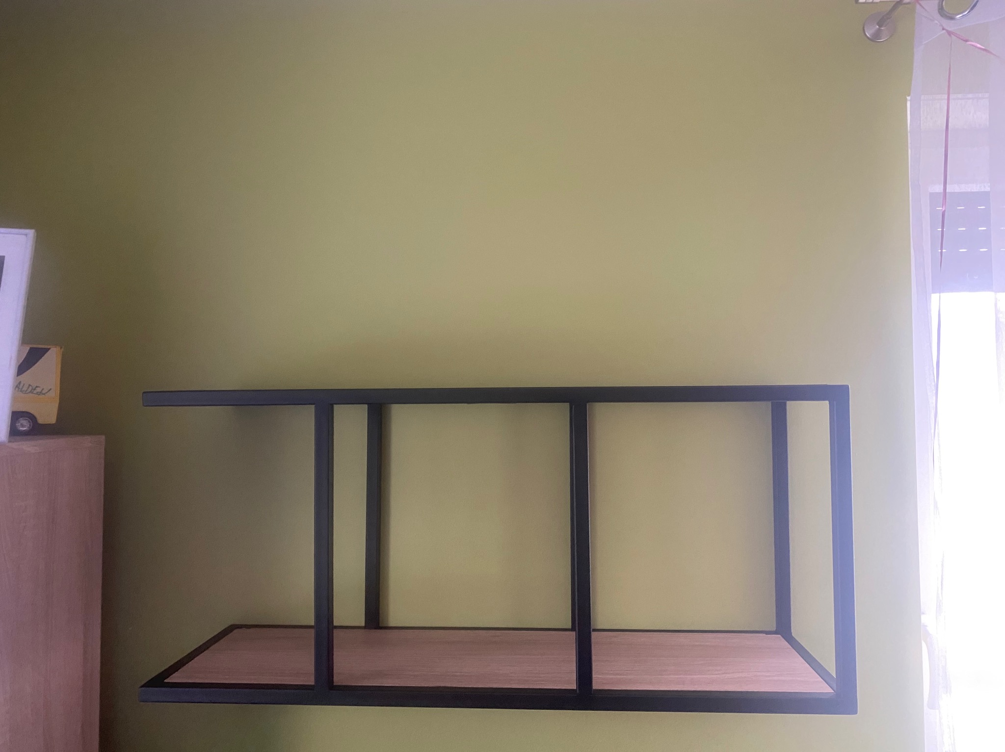 Półka wisząca, metalowa rama w kolorze czarnym, drewniane półki na tle zielonej ściany, widoczne fragmenty szafki i zasłony.