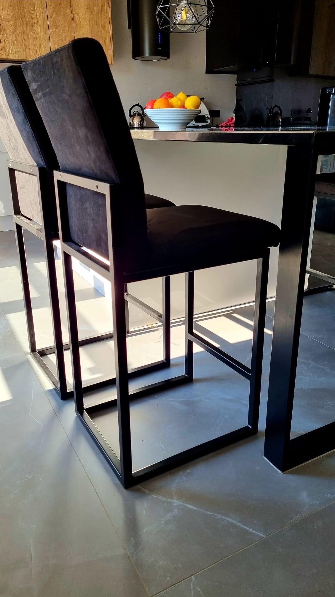 Dwa nowoczesne, czarne hokery z metalową ramą i welurowym siedziskiem, ustawione przy blacie kuchennym, oświetlone promieniami słonecznymi padającymi na szarą, marmurkową podłogę.