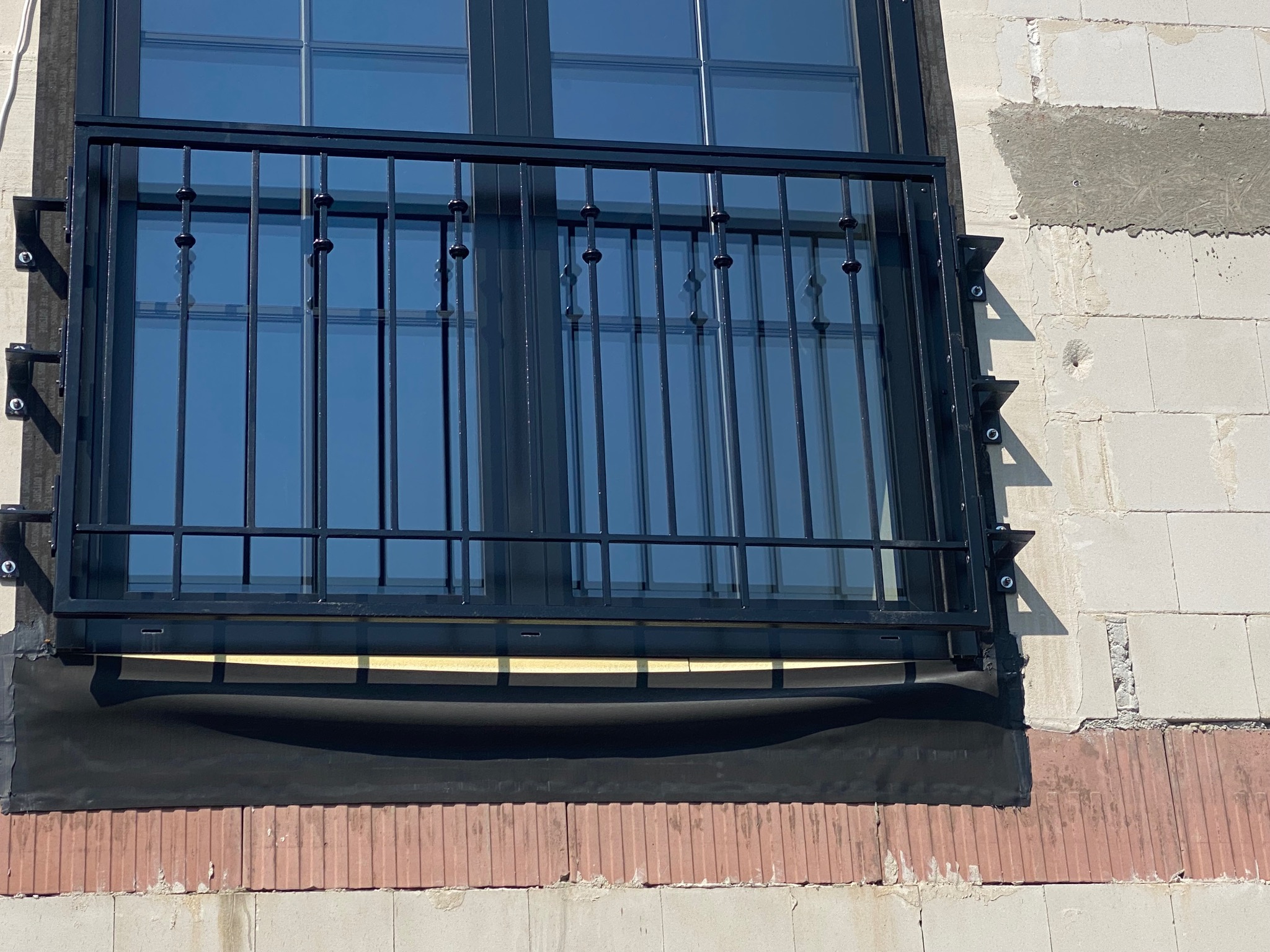 Czarne, metalowe zabezpieczenie okienne z prętami, zamontowane na ścianie z białych bloczków i czerwonej cegły. Widoczne mocowania i fragment okna.