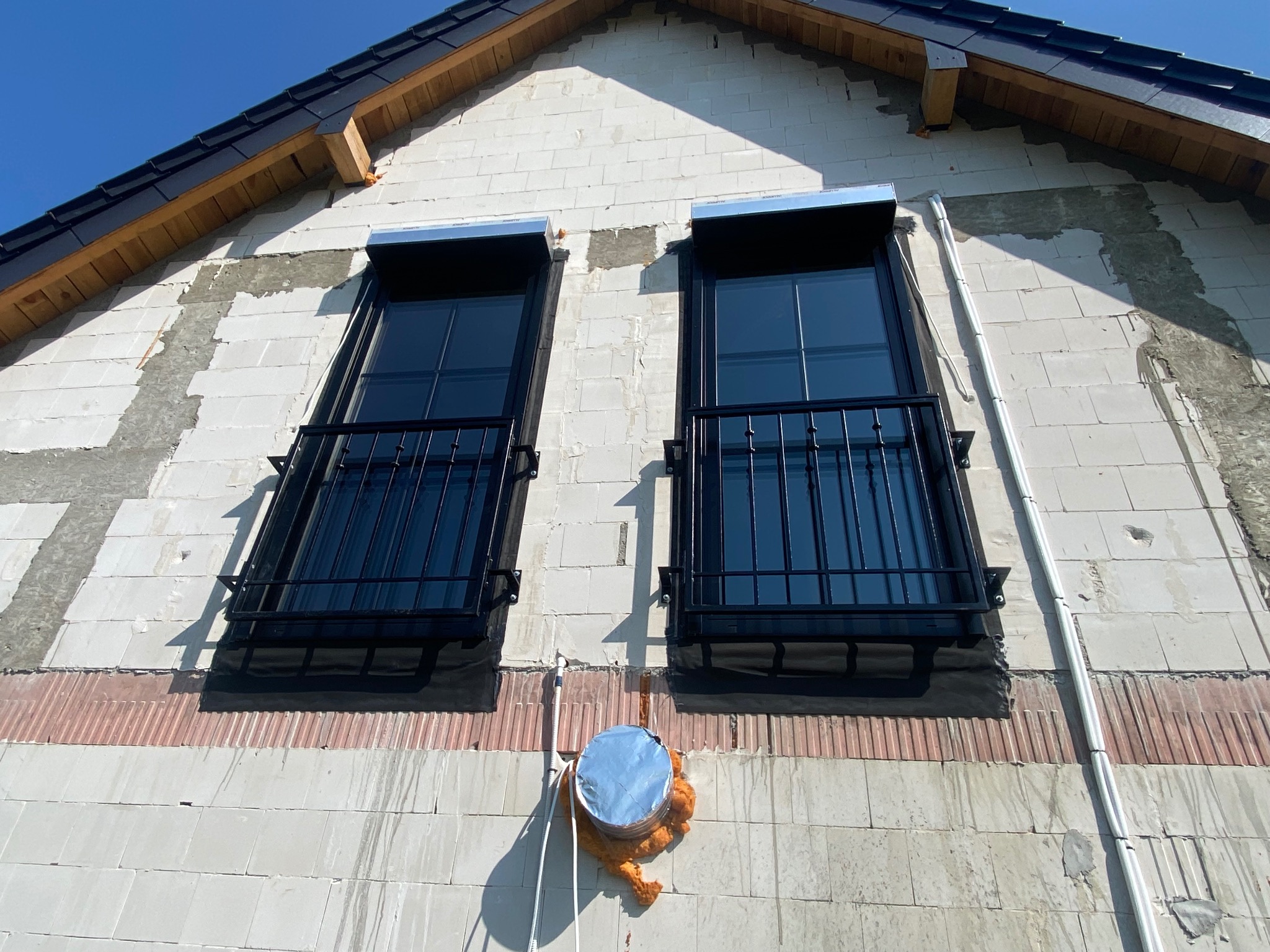 Dwa okna z czarnymi, metalowymi balustradami na surowej elewacji budynku z białego bloczka, widok z dołu, w tle błękitne niebo.