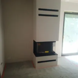 Wnętrze w trakcie wykańczania z nowoczesnym, białym kominkiem z poziomymi wnękami, surowe ściany i podłoga z widocznymi śladami prac, okno balkonowe w tle.
