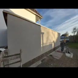 Pracownik w kombinezonie z kielnią w ręku, przygotowuje się do nałożenia zaprawy na elewację budynku, widoczne rusztowanie i zabezpieczenia na podłożu.