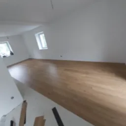 Jasny pokój na poddaszu z nowo ułożoną, drewnopodobną podłogą panelową; widoczne niedocięte panele i listwy przypodłogowe.