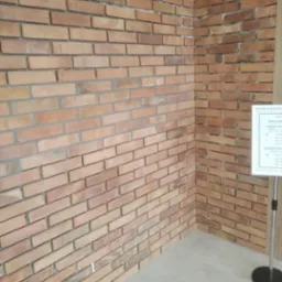 Wewnętrzny narożnik ściany wyłożonej cegłą klinkierową w odcieniach czerwieni i brązu, z widoczną fugą, obok tablicy informacyjnej na stojaku.