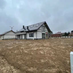 Surowy stan domu murowanego z czarną dachówką i oknami, widoczne elementy szalunku, teren wokół wyrównany, dzień pochmurny.