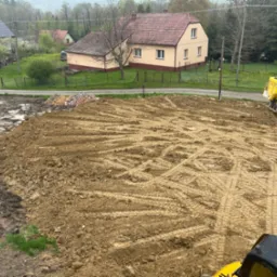 Przygotowany teren pod budowę z widocznym żółtym wywrotką i fragmentem koparki, ślady opon na ubitej ziemi, w tle zabudowa wiejska i drzewa.